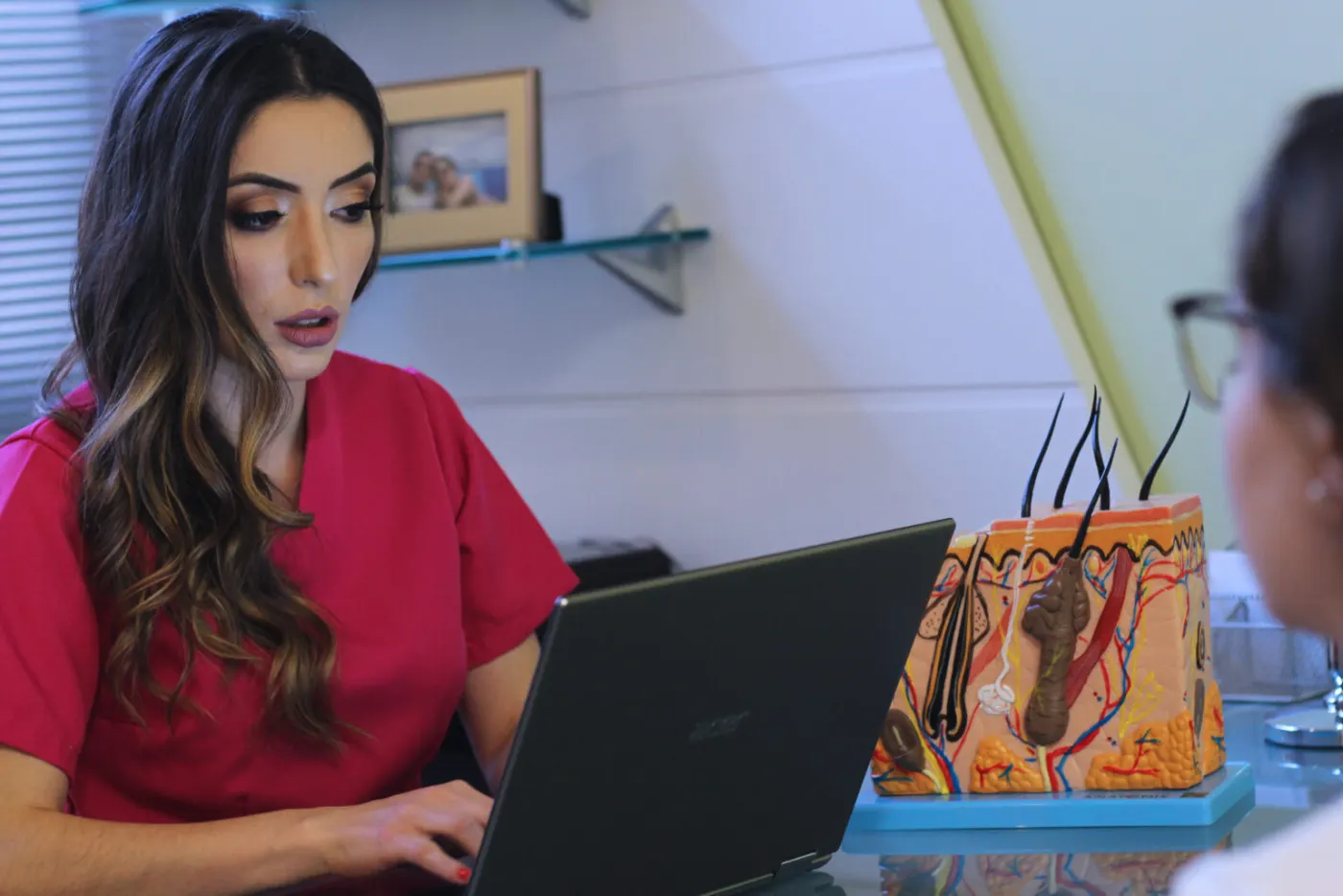 Dra. Monique Rosário em seu consultório, com laptop e um modelo anatômico de pele sobre a mesa