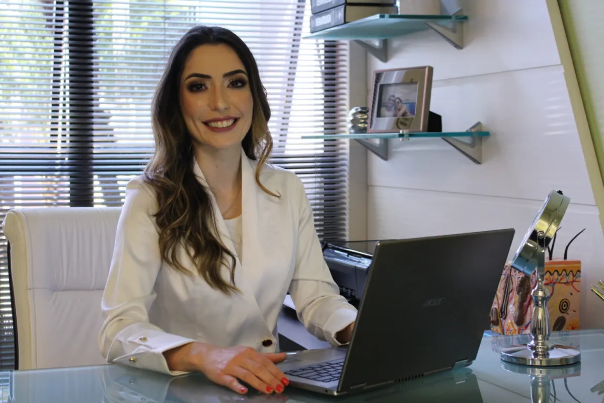 Dra. Monique Rosário sorrindo ao laptop em seu consultório, pronta para a teleconsulta